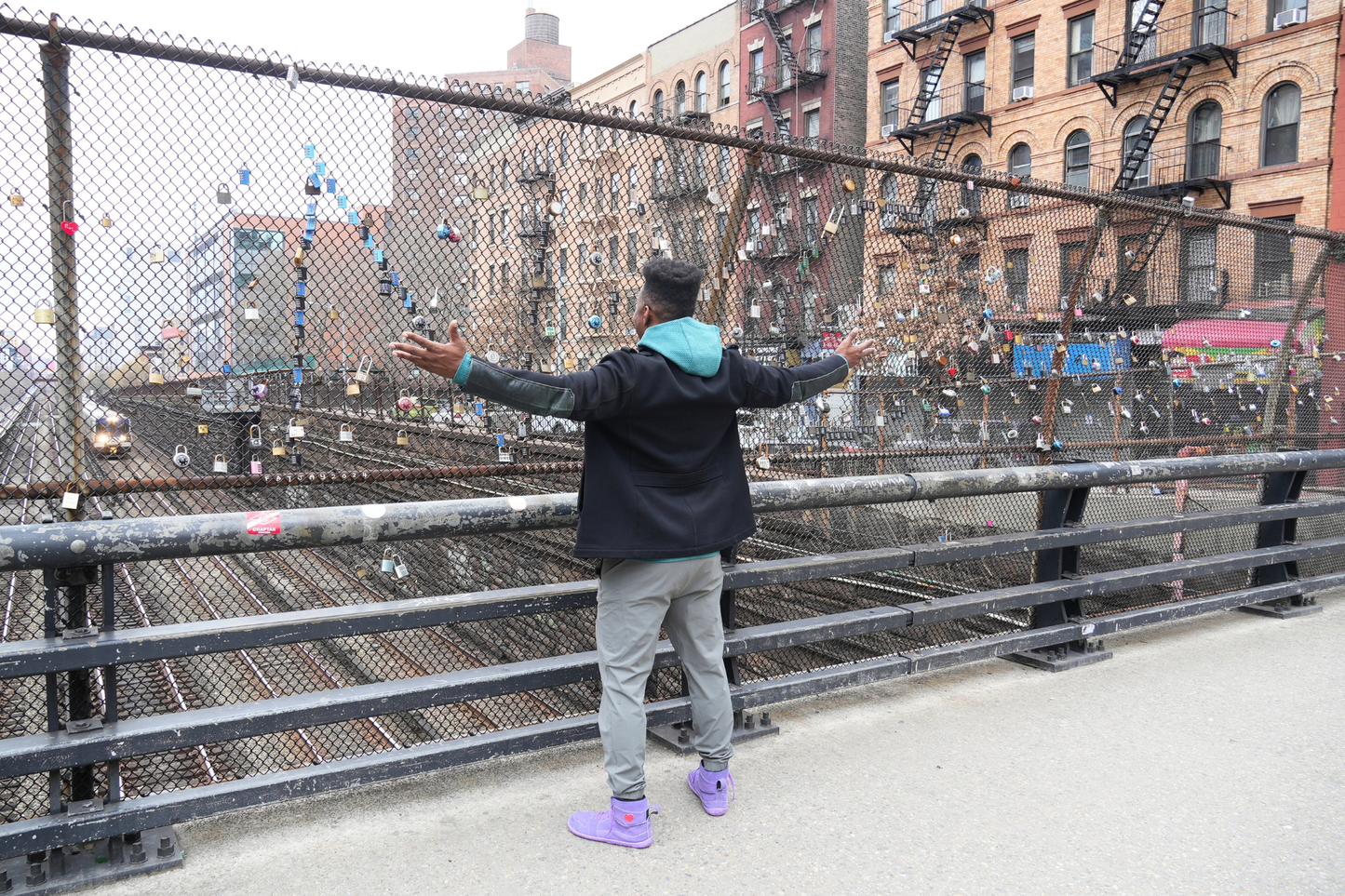 Person standing on a bridge with arms outstretched, looking at padlocks on a fence.