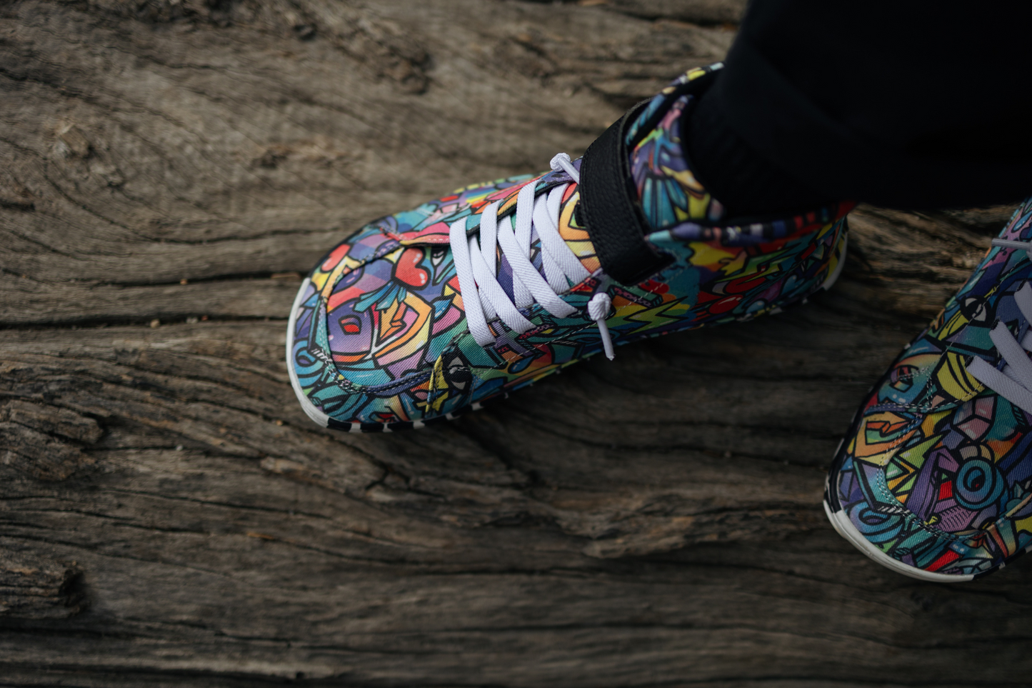 PaperKrane foot-shaped Mid Top barefoot shoes with graffiti print on cotton, black velcro strap, and zebra soles. Another view from above the shoes, this time standing on a log with one foot in front of the other. The person is wearing black socks and pants.