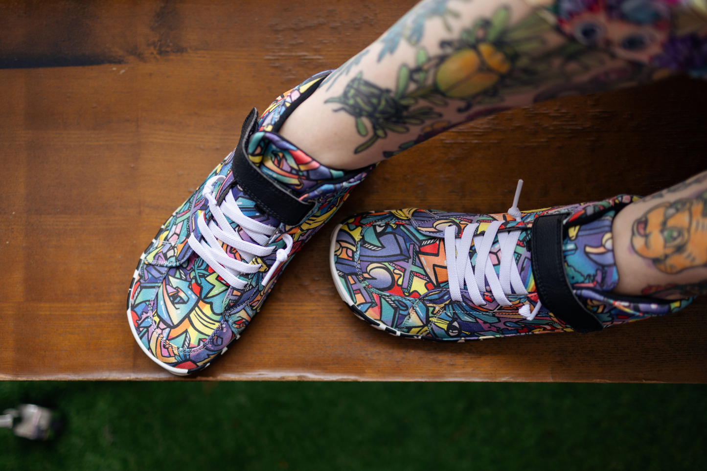 PaperKrane foot-shaped Mid Top barefoot shoes with graffiti print on cotton, black velcro strap, and zebra soles. The shoes can be seen from above on an adult's tattooed legs. They're sitting on a brown wooden bench.