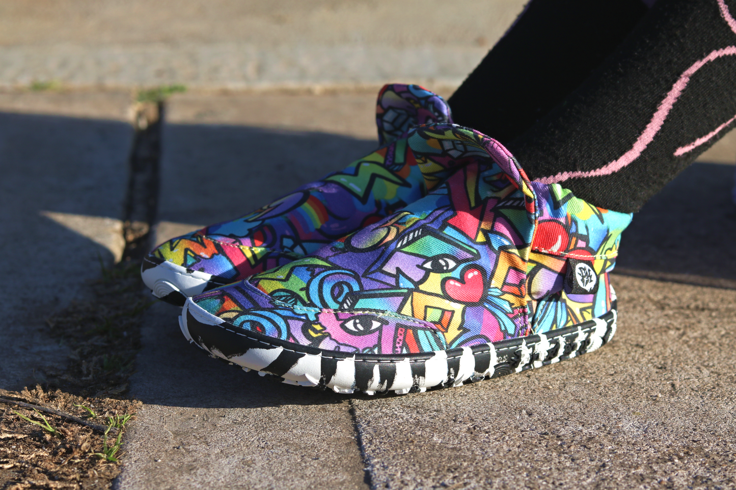 PaperKrane Graffiti signature style barefoot shoe slip ons, with hand-drawn graffiti print and zebra soles, on the feet of a child who is sitting on the footath. The shoe has a pop up tongue, and elastic ankles.