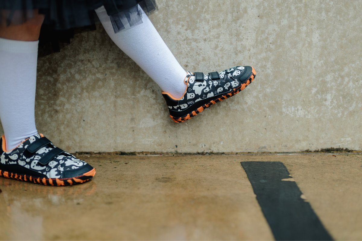 A person wearing black and white ghost-print velcro sneakers with bright orange tiger soles, paired with white knee-high socks, standing on a wet concrete floor against a textured wall.