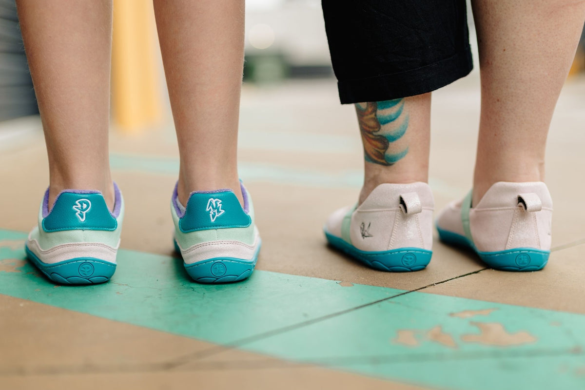 Two pairs of PaperKrane barefoot shoe styles shown from the back, worn by people standing on a painted green pavement line. The shoes feature a glittery pastel pink, mint, and teal upper with a bright teal heel tab and sole, plus light purple side accents on the sneaker.