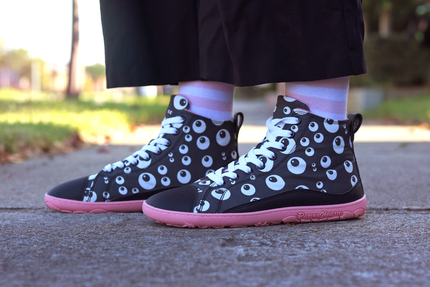 PaperKrane barefoot eyeball boots on legs with stripey socks and black pants, on a footpath. The boots are black with cartoon eyeballs, elastic laces, they're a foot-shaped, zero-drop design, on a salmon coloured sole.