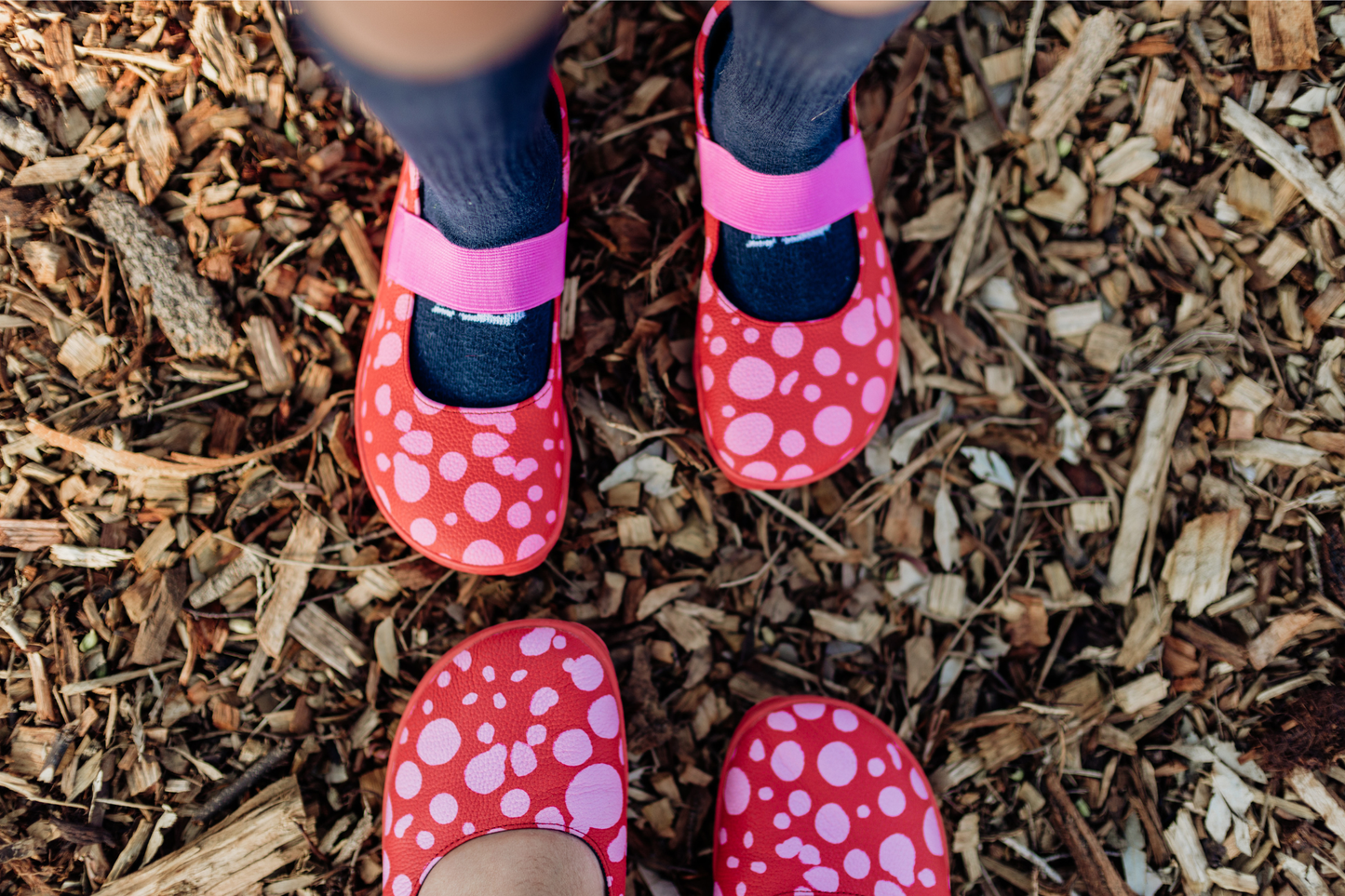 Red leather foot-shaped mary jane shoes on kid and adult feet, with pink dots, and pink straps on a ground covered with wood chips.