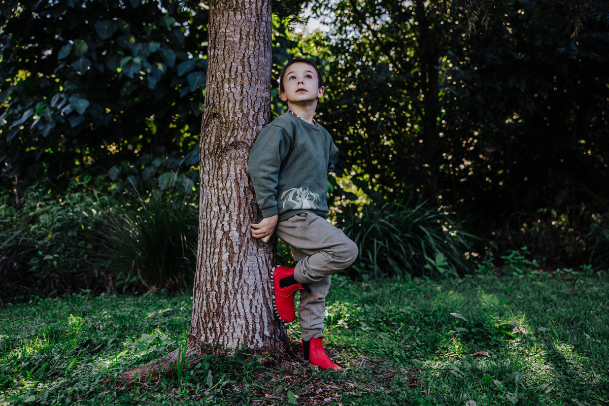 A young boy stands leaning against a tree trunk in a grassy, wooded outdoor setting. He is wearing a dark green long-sleeve shirt, beige cargo pants, and bright red leather PaperKrane chelsea boots.