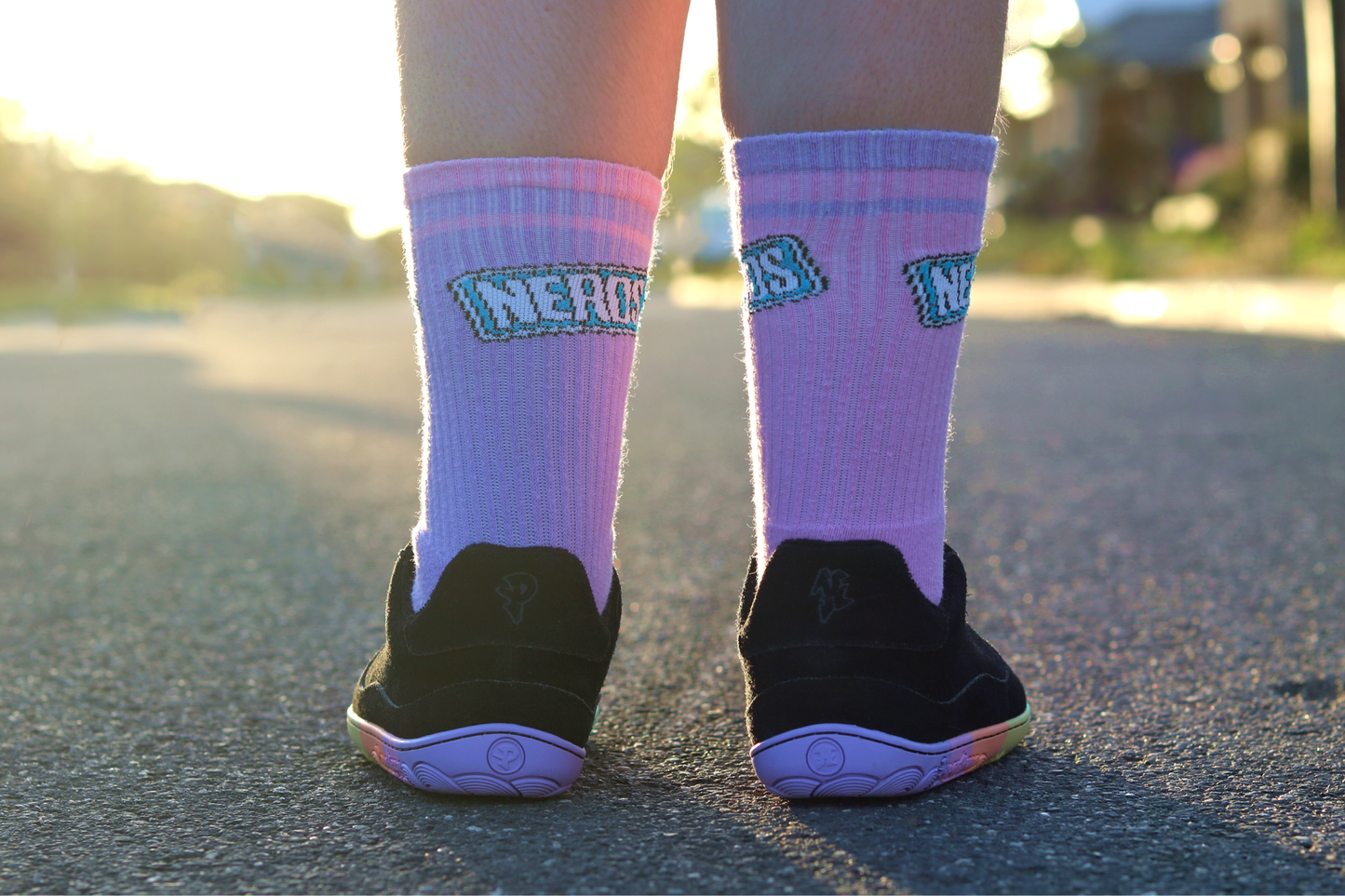 Person wearing black suede sneakers with pink socks with 'NERD' text, standing on a road.