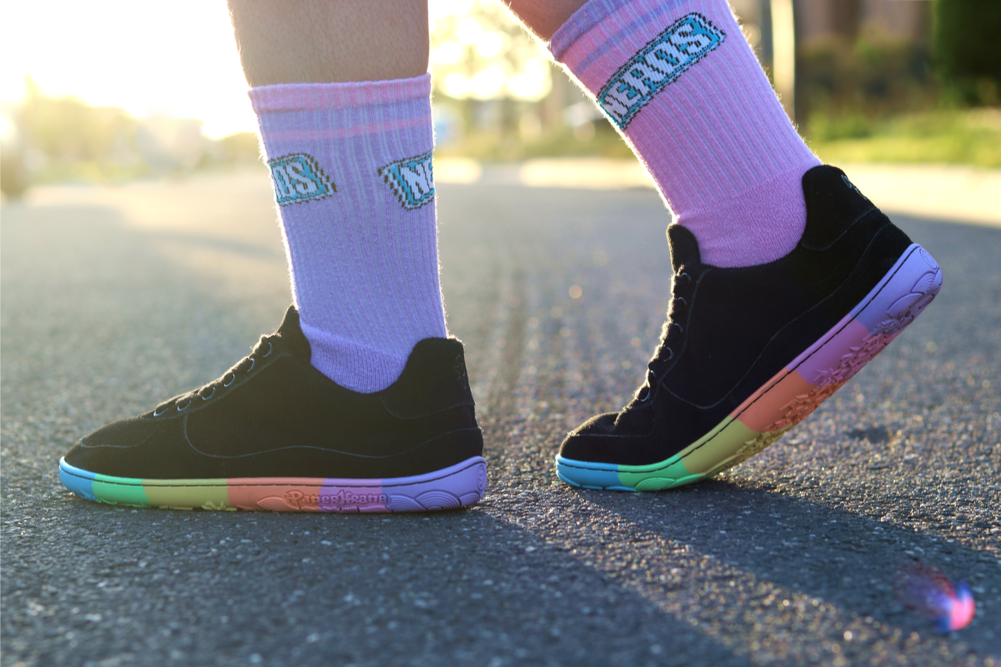 Person wearing black sneakers with rainbow soles and pink socks with text on a blurred outdoor background