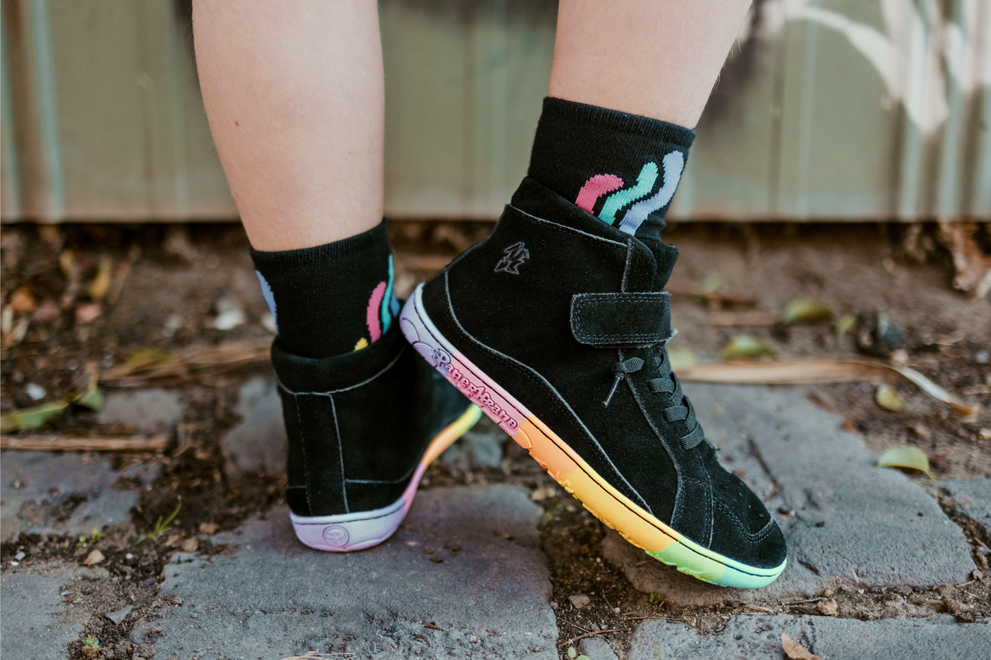 Person wearing black mid-top sneakers with colourful sole and socks with abstract designs on a concrete surface.