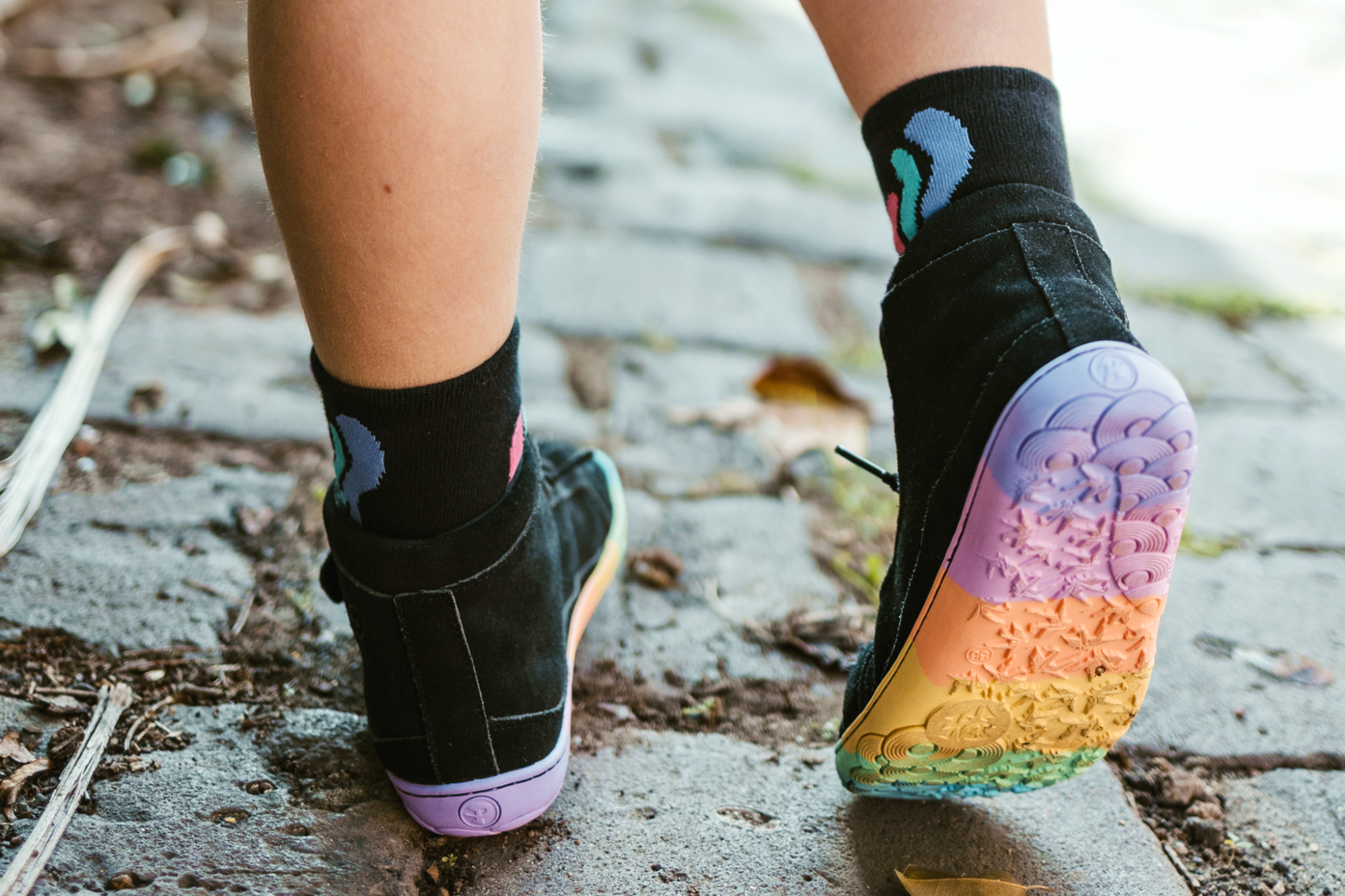 Person wearing black socks with colorful designs and rainbow-coloured shoes on a concrete surface.