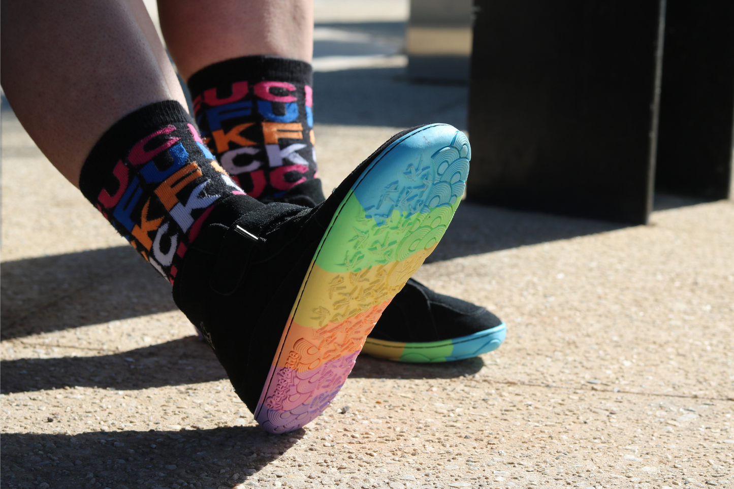 Person wearing colourful socks with letters and a rainbow-patterned shoe on a concrete surface.