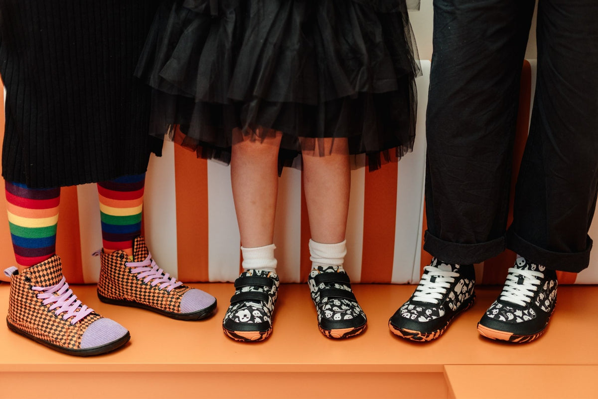 Three people standing on an orange platform, showing the lower half of their outfits. The center person wears a black ruffled skirt and black-and-white ghost patterned shoes with white socks. The lace up version of these ghost shoes is to the right, the person wears black trousers, and to the left, a person in rainbow socks and a sweater dress is wearing the orange argyle tall boots with their purple corduroy toes and elastic laces.
