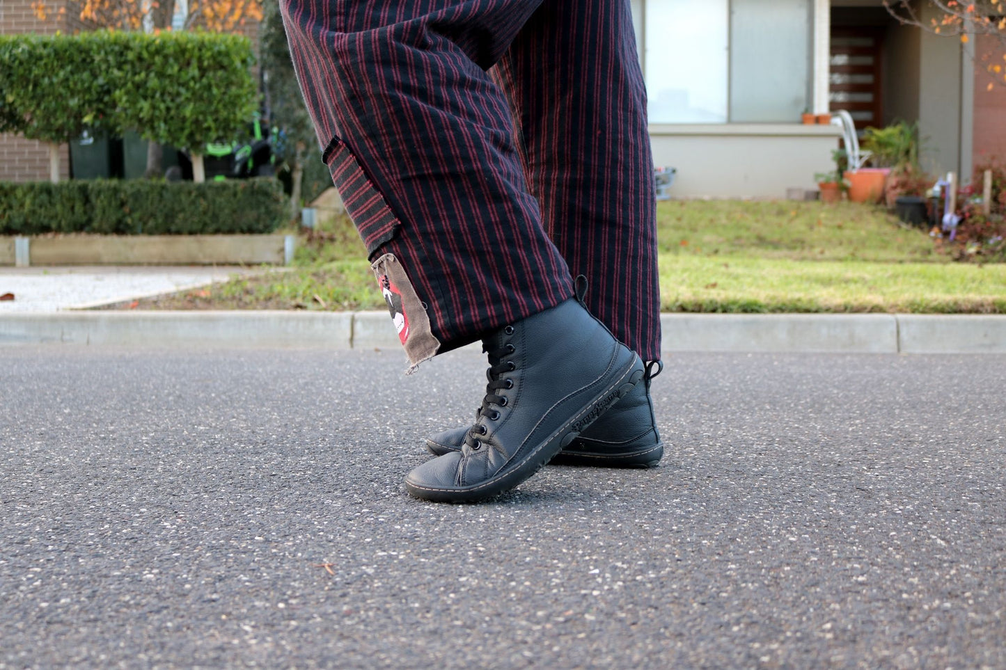 A person wearing black slip on boots with elastic laces and dark red-and-black vertically striped pants, standing on asphalt outdoors with a house and lawn in the background.