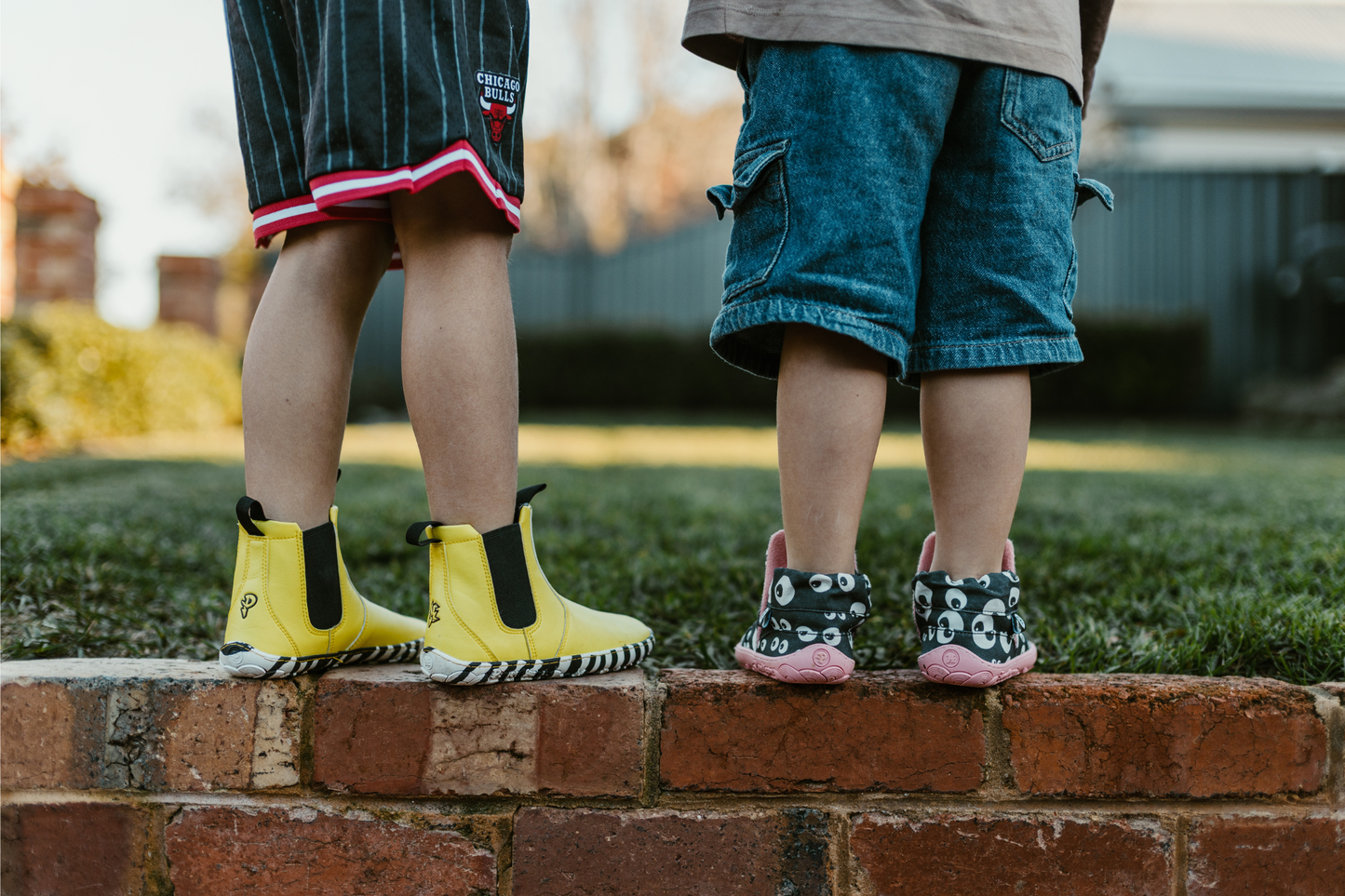 Two children standing on a brick ledge outdoors, shown from the back. One child wears bright yellow chelsea boots with black trim and zebra soles, and the other wears patterned black cotton slip-ons with eyeball print and salmon pink soles and lining.