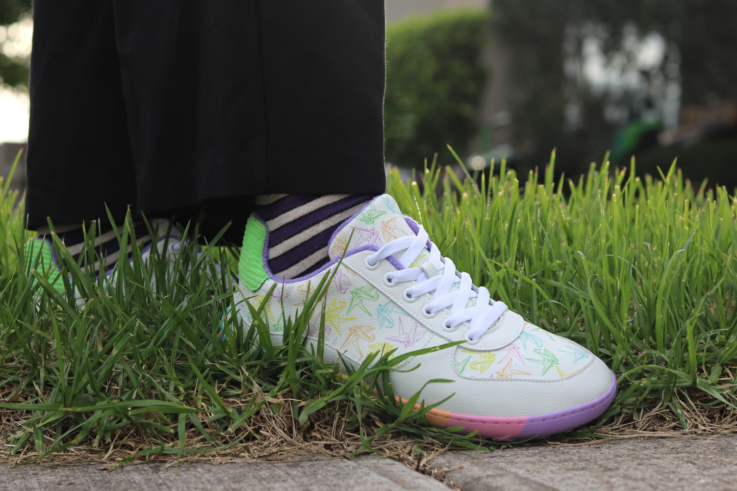 White barefoot sneakers from PaperKrane with a zero drop rainbow sole, and rainbow origami cranes printed on them. A person stands on grass with purple striped merino socks.