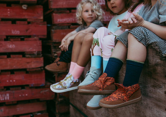 Four kids sitting on a wooden box with different coloured PaperKrane shoes on their feet.