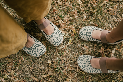 Two pairs of feet (adult and child) wearing cheetah patterned foot-shaped shoes standing on grassy ground with fallen leaves.