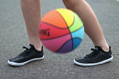 Person wearing black PaperKrane AKTIVE sneakers with a colourful basketball on a gray surface