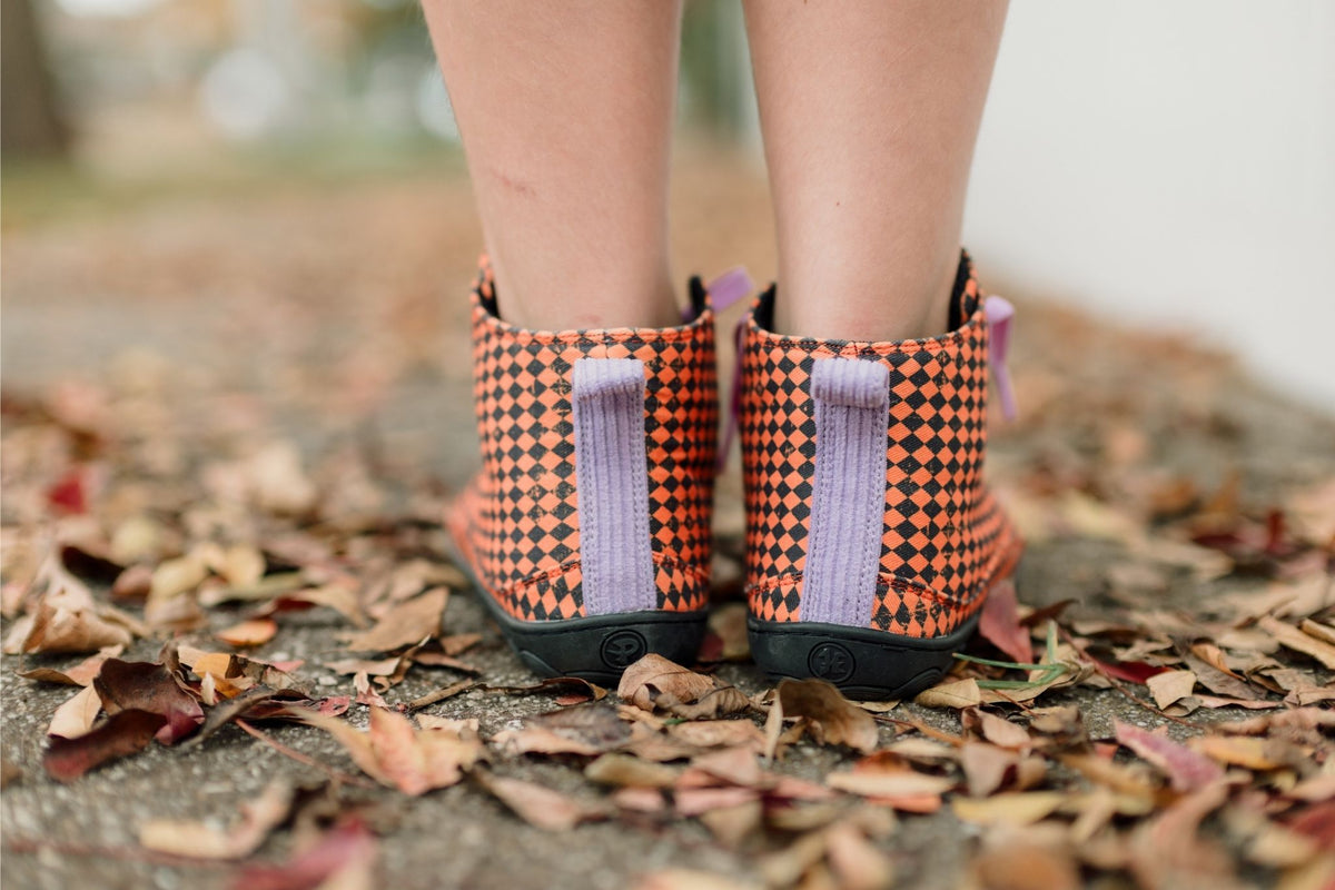 A close-up of a person's legs wearing orange and black argyle tall boots from PaperKrane with purple corduroy details, standing on a stone path covered in fallen autumn leaves.