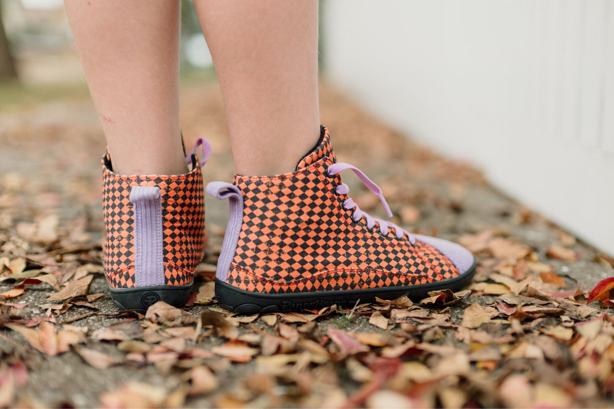 A close-up of a person's legs wearing orange and black argyle print high-top PaperKrane sneakers with lavender laces and corduroy details, standing on a ground covered in dry autumn leaves.