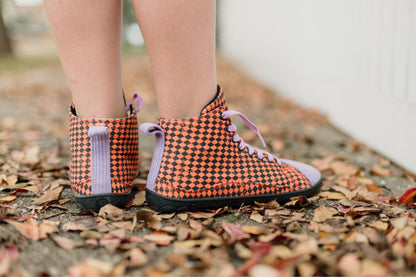 A close-up of a person's legs wearing orange and black argyle print high-top PaperKrane sneakers with lavender laces and corduroy details, standing on a ground covered in dry autumn leaves.