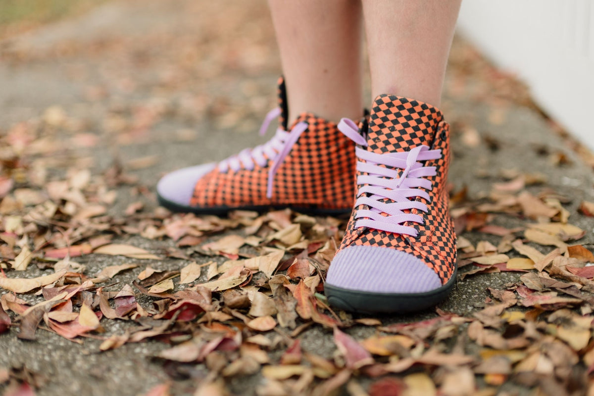 A close-up of a person's legs wearing orange and black checkered high-top sneakers with light purple laces and purple corduroy toe caps, standing on a concrete path covered with dry autumn leaves.