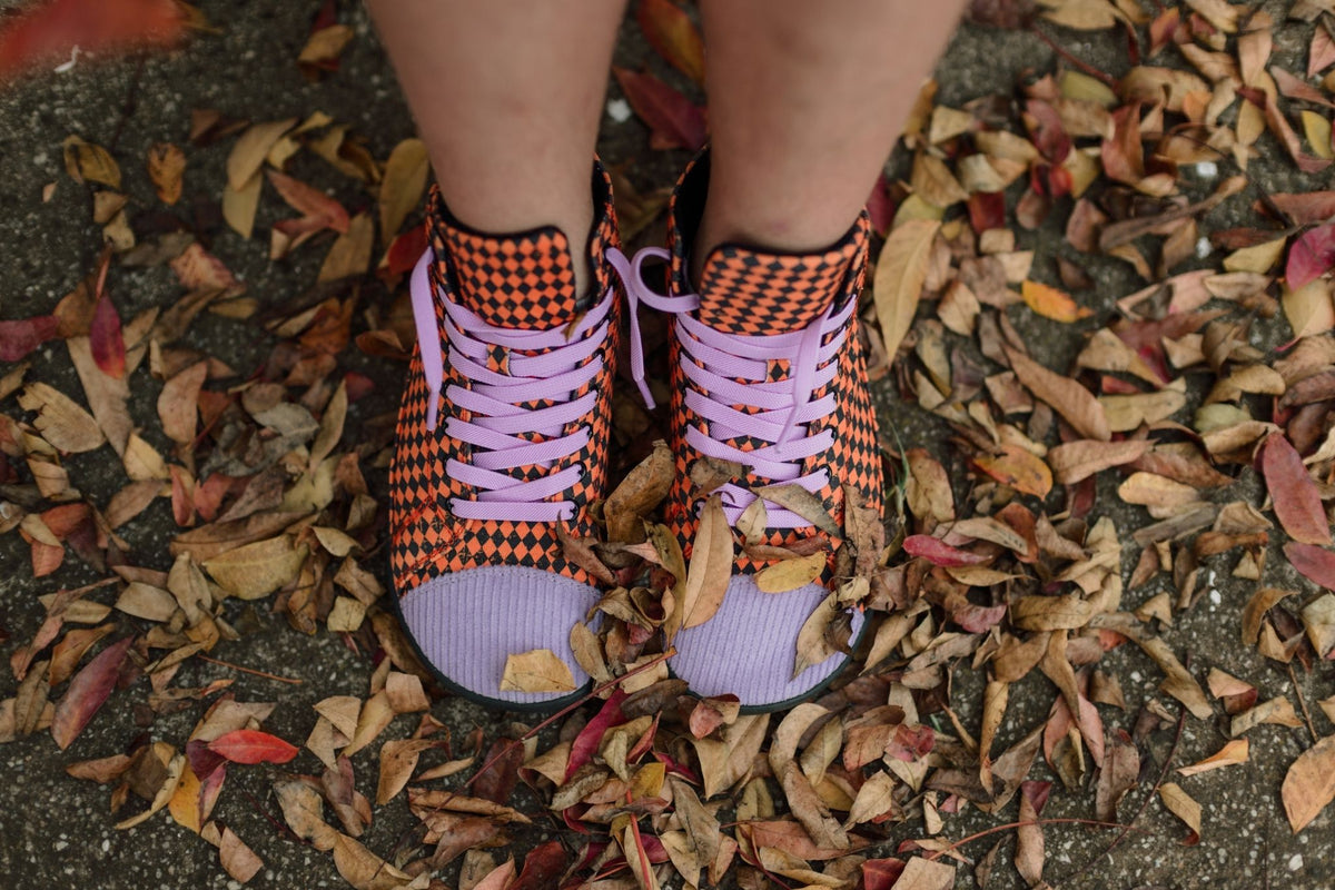 A person wearing orange and black checkered high-top sneakers with lavender laces and lavender corduroy toe caps, standing outdoors on a ground covered in dry autumn leaves.