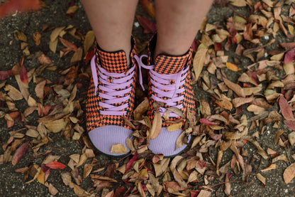 A person wearing orange and black checkered high-top sneakers with lavender laces and lavender corduroy toe caps, standing outdoors on a ground covered in dry autumn leaves.