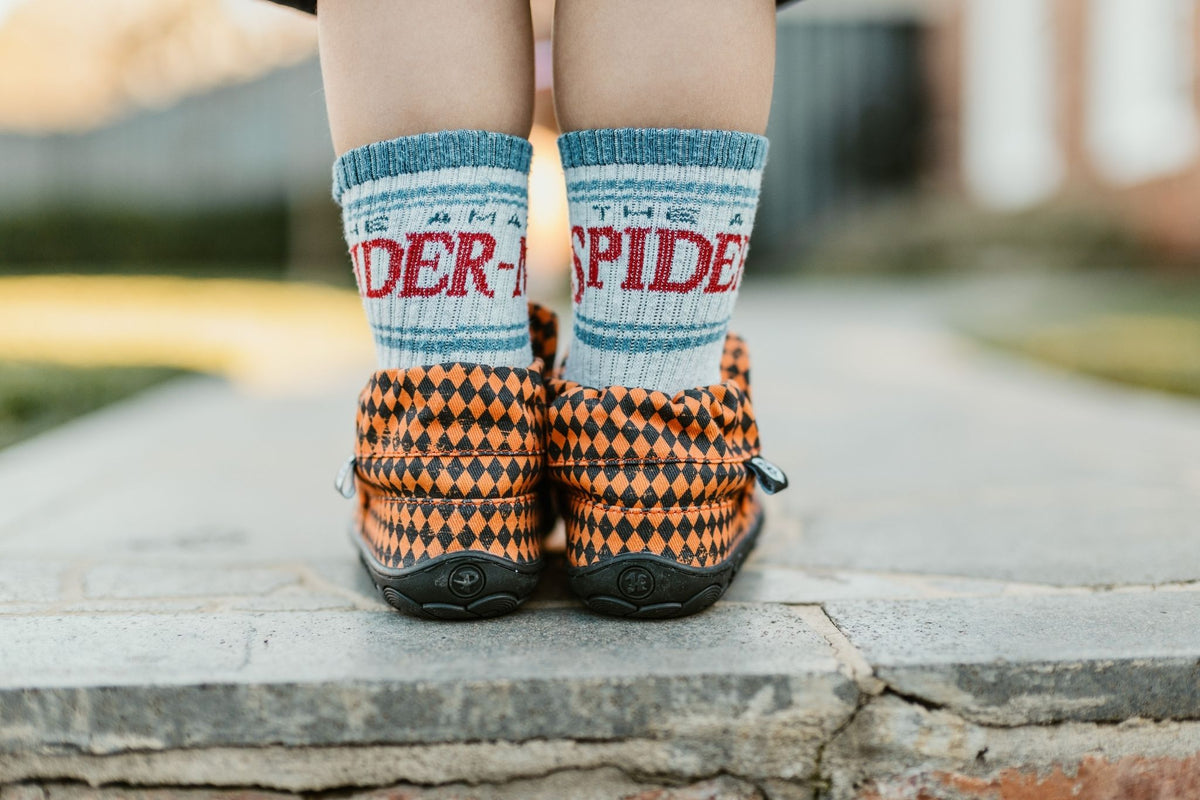 A close-up lifestyle shot of a child's legs standing on stone steps, wearing orange and black harlequin-patterned slip-on shoes with blue and white socks featuring 'THE AMAZING SPIDER-MAN' text.