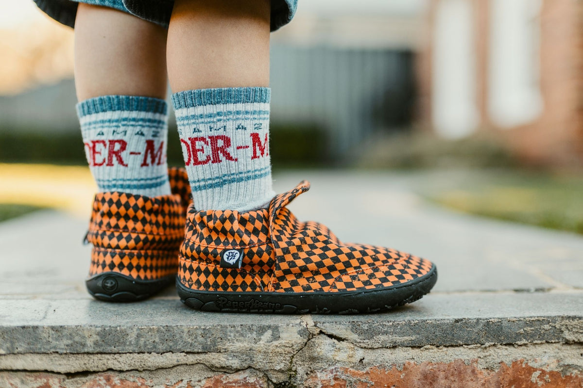 A close-up of a child's feet wearing orange and black checkered slip-on barefoot shoes from PaperKrane and blue-and-white socks with red lettering, standing on a stone ledge outdoors.