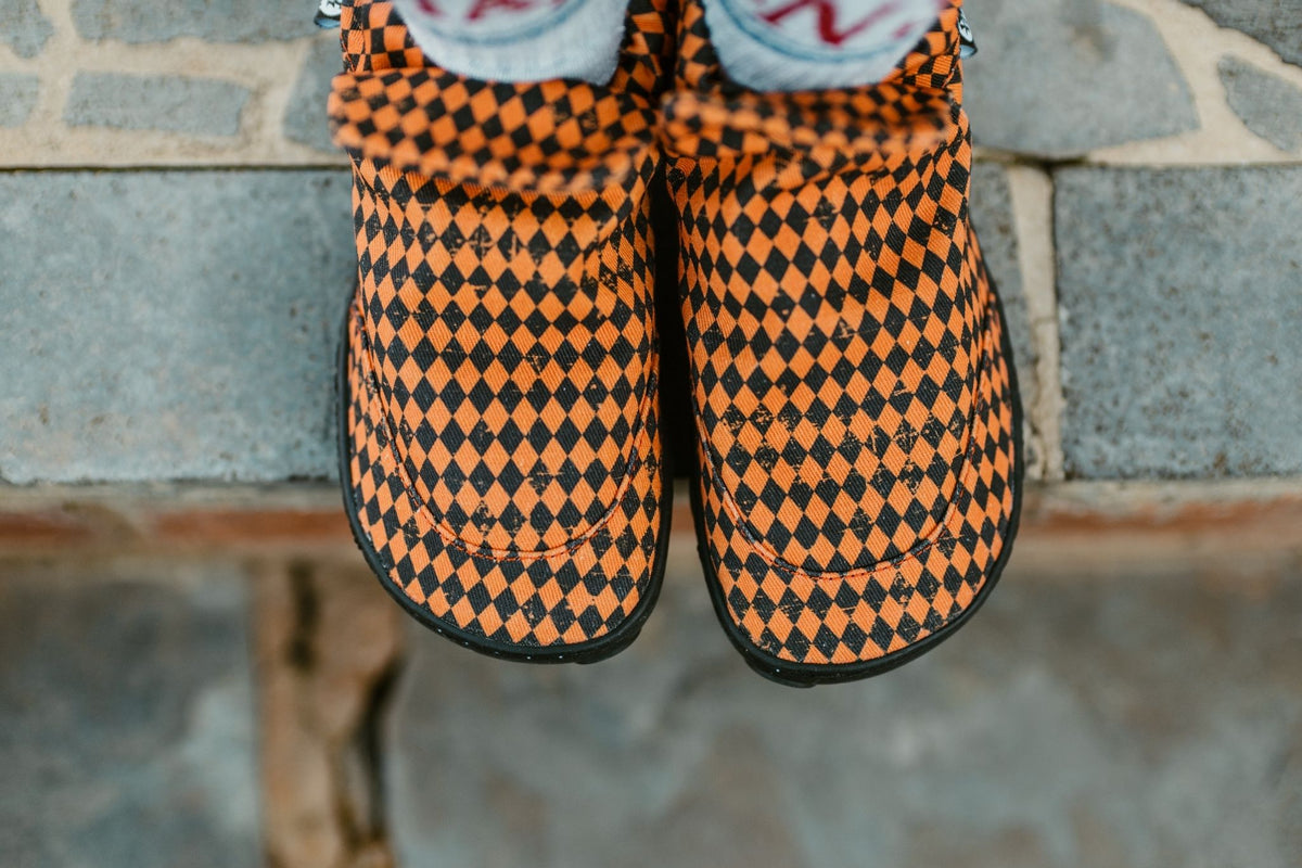 A close-up of a pair of slip-on canvas boots with a black and orange diamond argyle pattern, worn by a child standing on the edge of some stone steps.