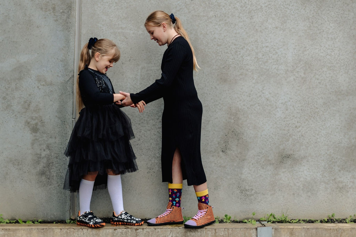 Two girls in black outfits standing against a gray concrete wall, holding hands. The girl on the left wears a black tulle dress, white knee-high socks, and black-and-white shoes, and on the right the girl wears the orange and black argyle tall sneakers with colourful mushroom socks.