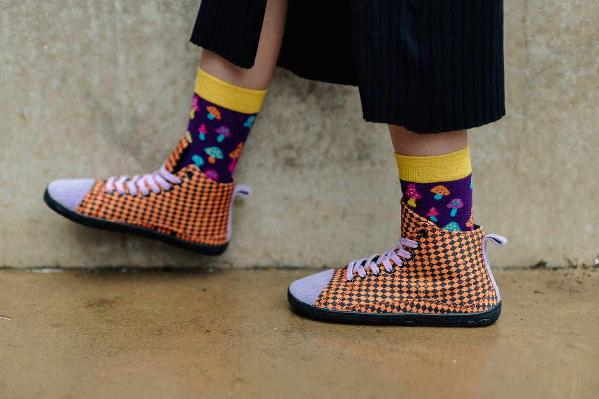 A person wearing orange and black argyle PaperKrane slip-on tall boots with light purple corduroy toes and heels, paired with purple socks featuring colourful mushroom prints and yellow cuffs, standing against a concrete wall.
