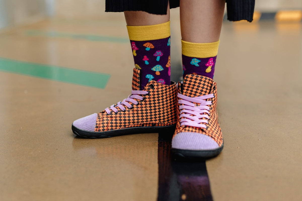 A person wearing orange and black argyle print sneakers with lavender corduroy toe caps and matching laces, paired with purple socks featuring a colourful mushroom pattern and yellow cuffs, standing on a we concrete surface.