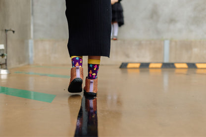 A person walking away on a concrete surface, wearing a long black ribbed skirt and colourful patterned socks with orange and black argyle checkered slip-on barefoot boots.