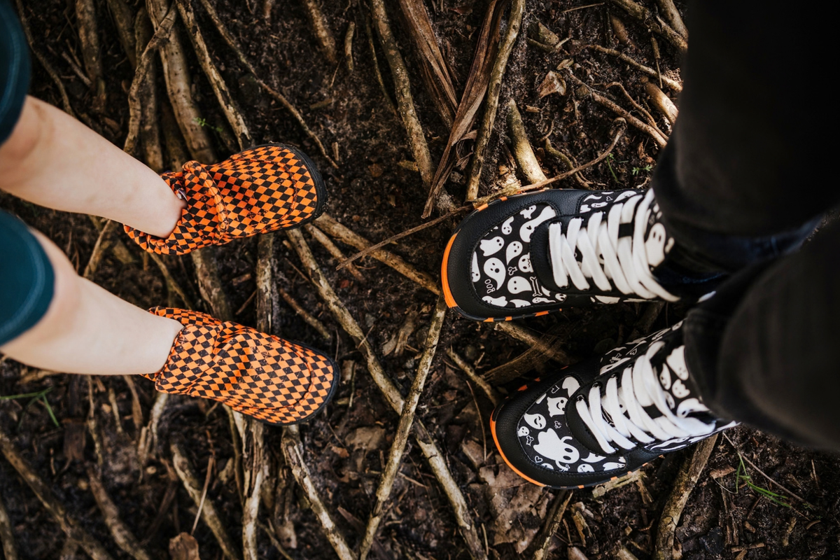 Children's legs in a pair of PaperKrane orange argyle print signature style slip ons is facing an adult who is wearing some foot-shaped PaperKrane sneakers with a black and white ghost print on them They have orange and black soles. They're standing on some we tree roots, and the view of the shoes is from above. 