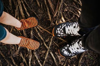 Children's legs in a pair of PaperKrane orange argyle print signature style slip ons is facing an adult who is wearing some foot-shaped PaperKrane sneakers with a black and white ghost print on them They have orange and black soles. They're standing on some we tree roots, and the view of the shoes is from above. 