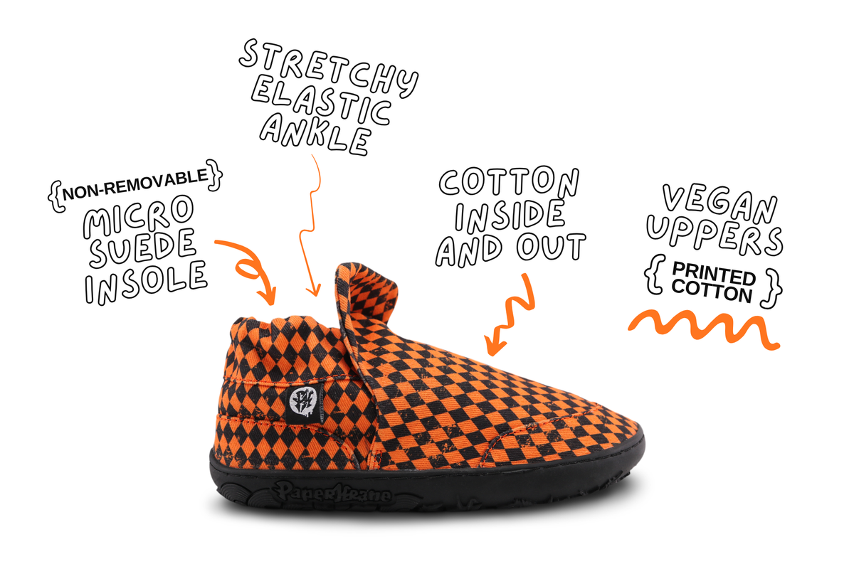 A single slip-on shoe with an orange and black checkerboard pattern, shown on a white background with handwritten callouts describing its features: stretchy elastic ankle, non-removable micro suede insole, cotton inside and out, and vegan uppers printed with cotton. These are the signature style barefoot shoes for kids from PaperKrane. 