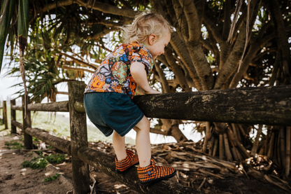 A small child wears a fun Halloween print tee, and some orange argyle print signature style PaperKrane slip on barefoot shoes. He's climbing on a fence near a big tree. 