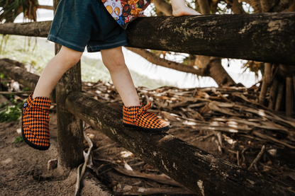 A small child wears some orange argyle print signature style PaperKrane slip on barefoot shoes while climbing on a fence near a big tree. 