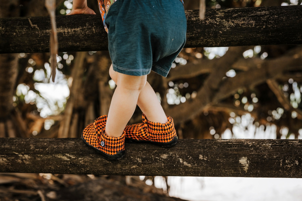 A small child wears some orange argyle print signature style PaperKrane slip on barefoot shoes. He's climbing on a fence near a big tree. 