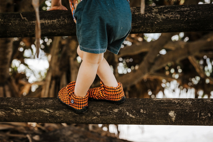 A small child wears some orange argyle print signature style PaperKrane slip on barefoot shoes. He's climbing on a fence near a big tree. 