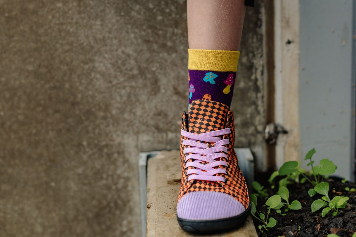 A close-up of a person's foot wearing a purple crew sock with colourful mushroom prints and an orange-and-black argyle barefoot tall sneaker with purple laces and toe detail, standing on a stone ledge next to small green plants.