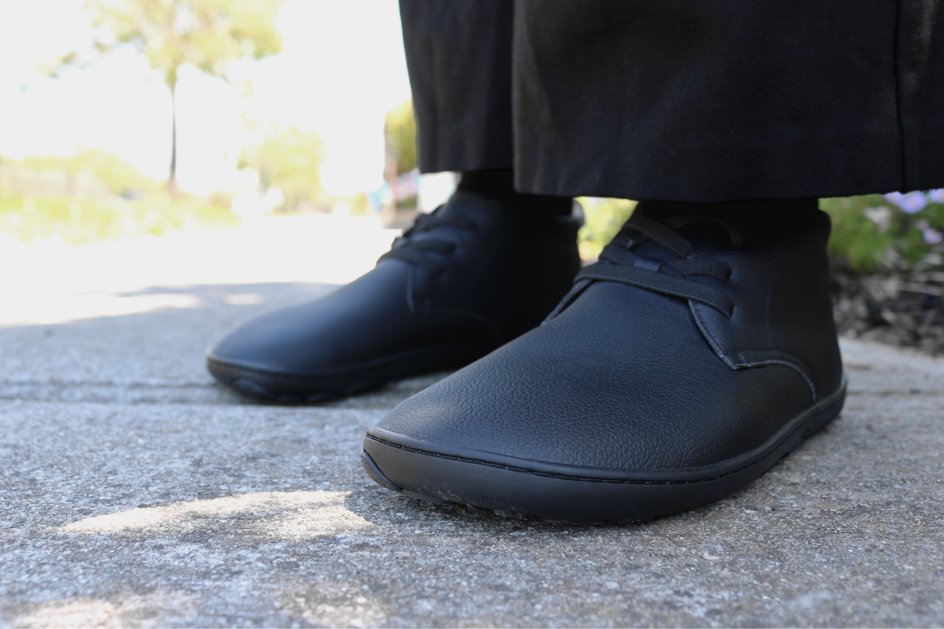 Person wearing black shoes on a concrete surface with blurred greenery in the background