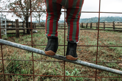 A child wearing red and green vertically striped leggings and black leather chelsea boots stands on a metal gate rail in an outdoor farm setting with fences and grass in the background.
