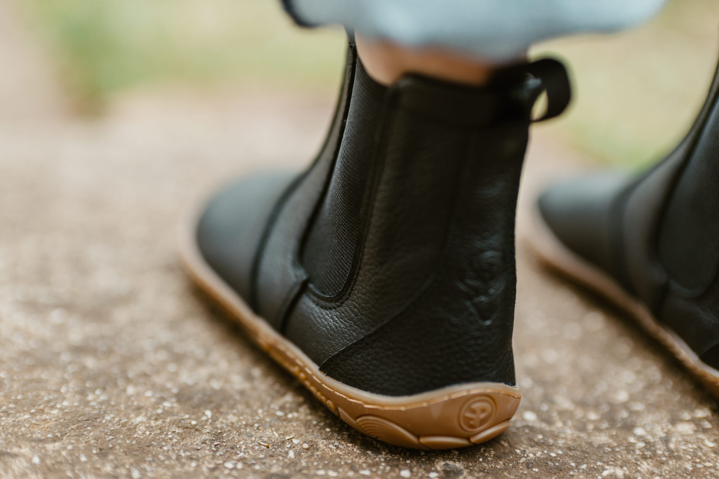 An adult wears a pair of black leather barefoot chelsea boots with PaperKrane logo on the sole. The leather is soft and tumbled giving it a natural look. The person is wearing jeans on a concrete path. This shows the back of the left boot with the embossed "P". 