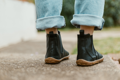 A person wearing folded up jeans is showing the back of a pair of black leather chelsea boots from PaperKrane. The boots are barefoot zero drop style with gum soles, on a concrete path. 