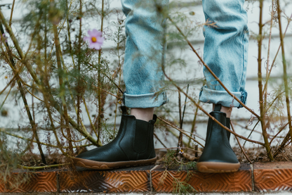 A person wearing PaperKrane black leather chelsea boots is standing on a ledge with some weedy looking plants around. You can see the foot-shaped toe box of the boot pointing towards the camera. 