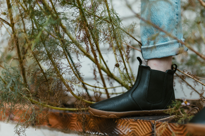 One leg of a person in jeans wearing PaperKrane barefoot black leather chelsea boots with a gum sole. Standing on a ledge with plants around them. 