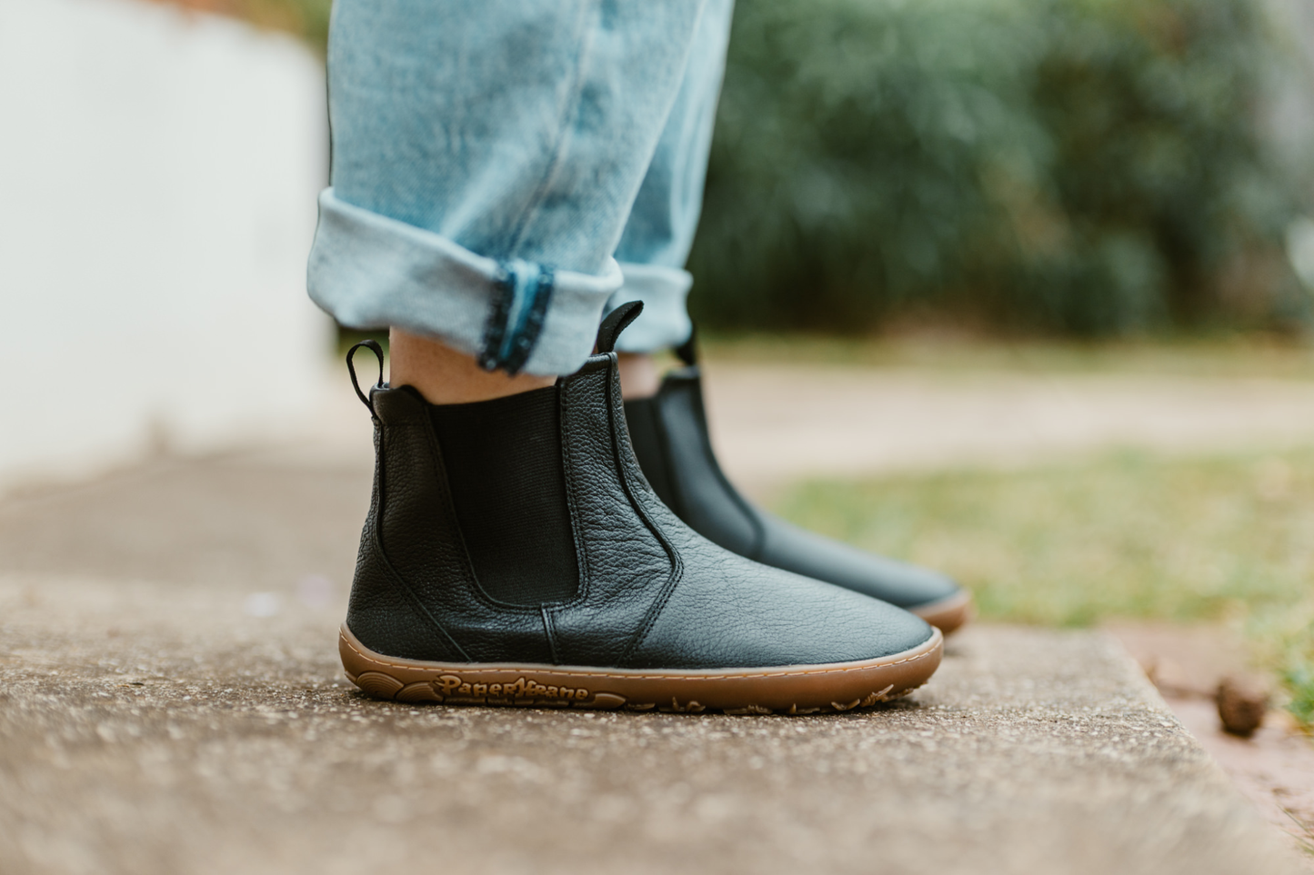 An adult wears a pair of black leather barefoot chelsea boots with PaperKrane logo on the sole. The leather is soft and tumbled giving it a natural look. The person is wearing jeans on a concrete path. 