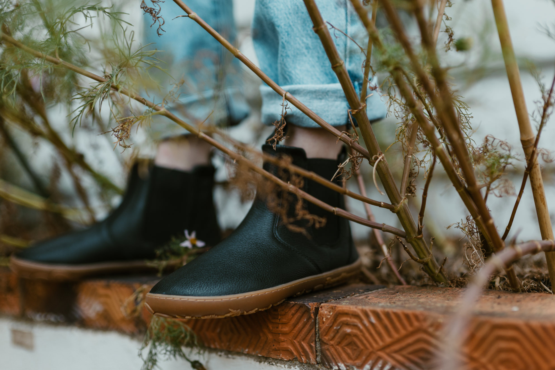 A person wearing PaperKrane black leather chelsea boots is standing on a ledge with some weedy looking plants around. 